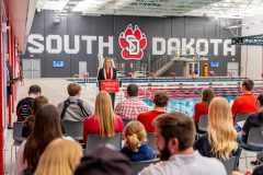 Woman stands at a podium and next to indoor pool speaking to a crowd.