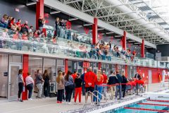 People standing around next to an indoor pool and seated in spectator seating.