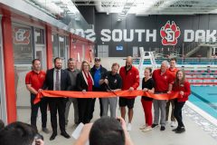 Eleven adults standing next to an indoor pool ceremoniously cut a large red ribbon. 